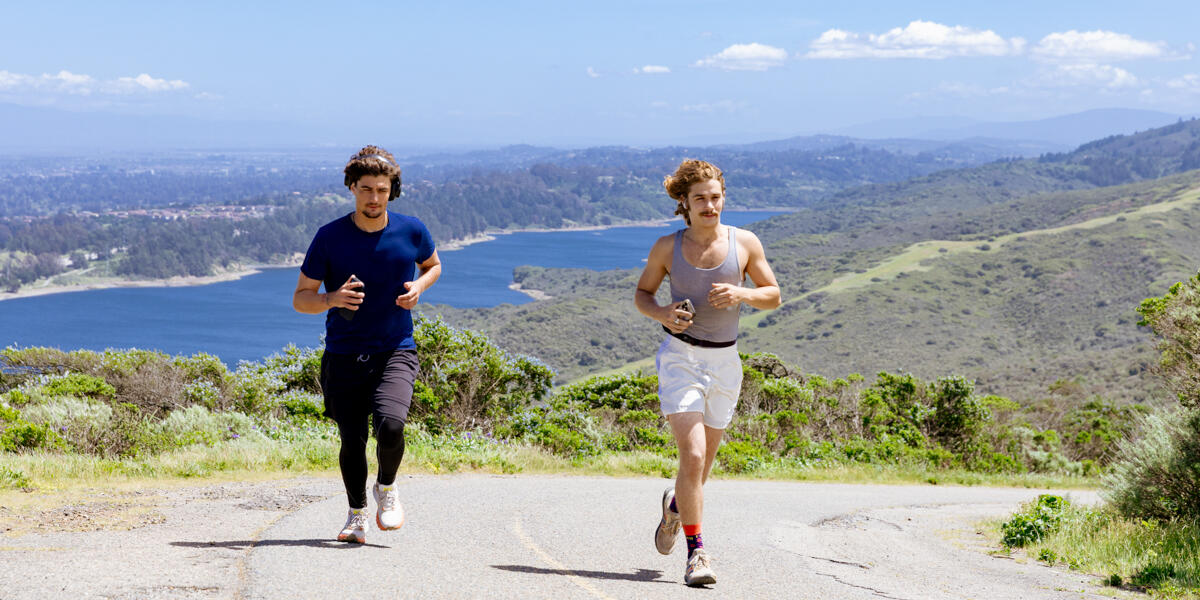 Two runners exercise on a paved trail at Sweeney Ridge, with a beautiful sunny backdrop of san andreas lake.