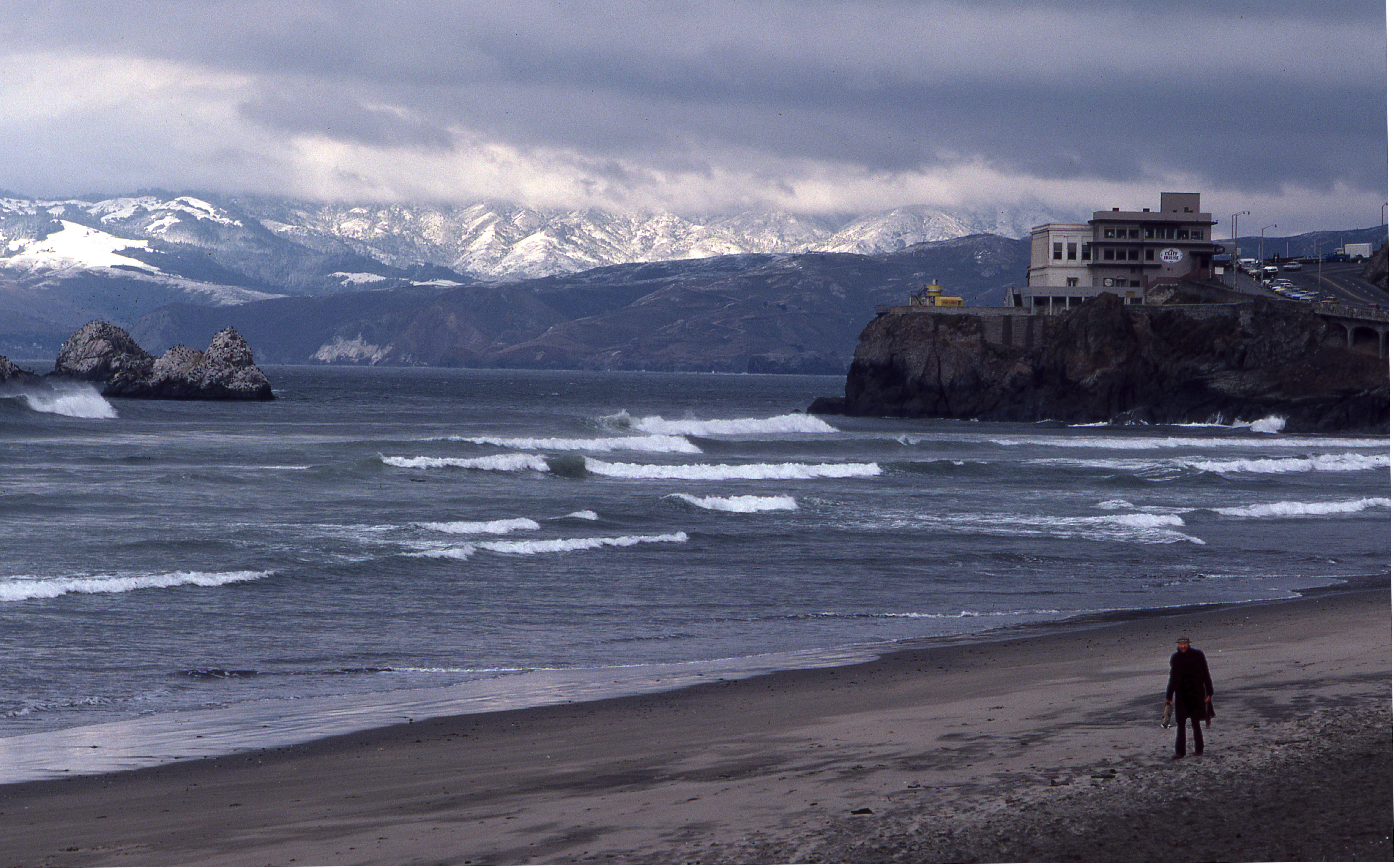 Person walking along Ocean Beach, in front of Cliff House, with snowy Marin Headlands in Background