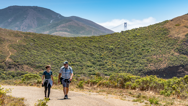 A couple hiking a dirt trail in the Marin Headlands. A Golden Gate Bridge tower can be seen peeking through the green mountain range in the background.