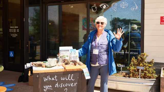 A park volunteer smiles and waves in front of a table of natural park items for visitors to explore.