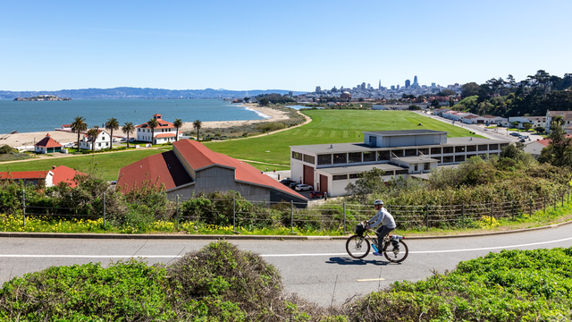 A cyclist is seen biking up a road with a vast backdrop of Crissy Field and the San Francisco skyline