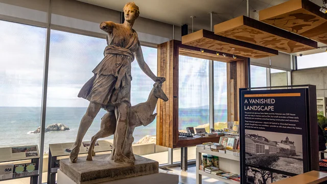 A view from inside the Lands End Lookout, showing a large stone sculpture of Diana, various signs and products, and a view of the pacific ocean through the windows.