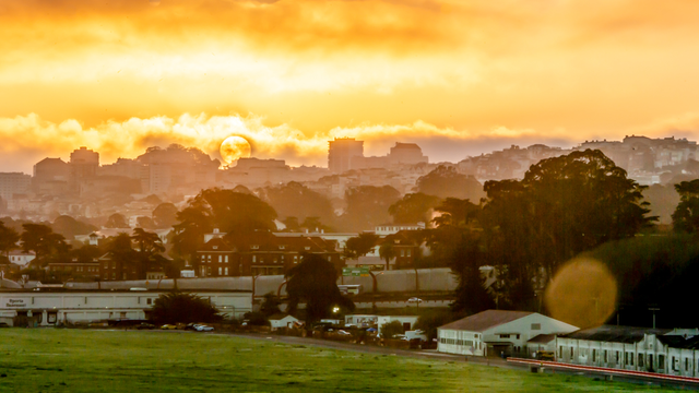 The bright yellow sun rises and shines through clouds over San Francisco skyline and Crissy Field.