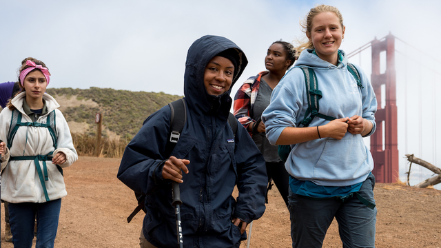 Four hikers trek down the SCA Trail in the Marin Headlands with the Golden Gate Bridge in the background.