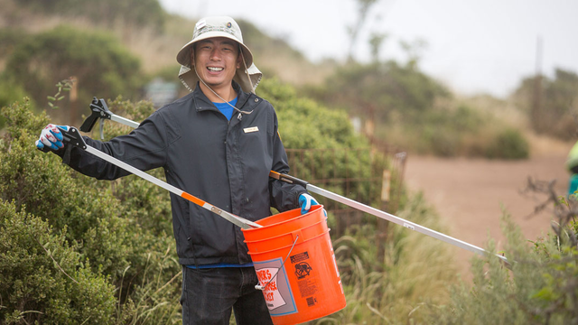 A volunteer holds a bucket and debris grabbing tool on a trail.