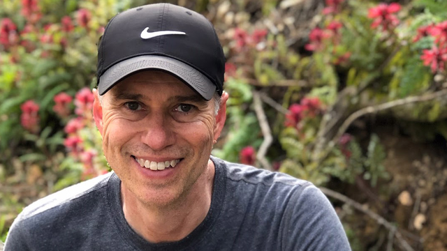 A volunteer smiles while seated in front of a blooming hedge
