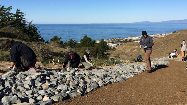 A small group of people focus on individual tasks while working outside on a trail overlook a body of water