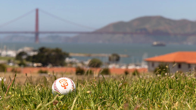 A baseball with the SF Giants logo lays on the green lawn at Fort Mason with a view of the Golden Gate Bridge in the background.