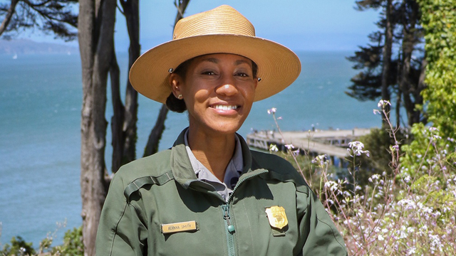 National Park Ranger Alanna Smith, a young woman, stands in the Presidio with the Pacific Ocean visible through the trees behind her.