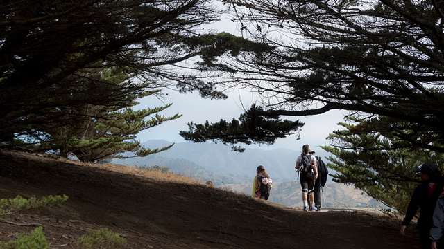 People hiking down a winding path through trees at Rancho Corral de Tierra, sloping hillsides are visible ahead and in the background.