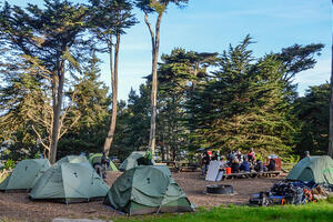 A group of adults sit outside on a sunny day eating lunch at a picnic bench under eucalyptus trees while surrounded by camping tents