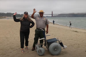 A Parks Conservancy staff member and a National Park Service staff member stand waving and smiling on East Beach next to a beach wheelchair.