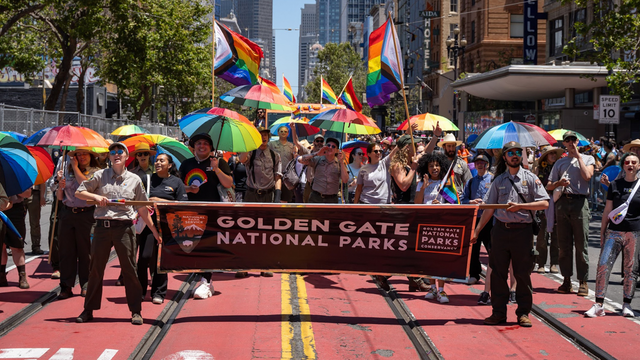 Smiling Parks Conservancy staff and NPS Rangers hold a Golden Gate National Parks banner and rainbow umbrellas in the Pride Parade.