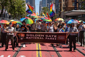 Smiling Parks Conservancy staff and NPS Rangers hold a Golden Gate National Parks banner and rainbow umbrellas in the Pride Parade.