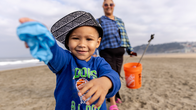 A happy child smiles while presenting a piece of collected trash to the camera during Coastal Clean Up Day at Ocean Beach