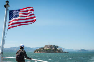 A park visitor looks toward Alcatraz Island from the ferry. An American flag waves in the breeze as they cruise over bay waters.