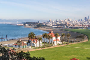 Aerial view of Crissy Field, buildings, the Warming Hut, palm trees, the golden gate, bay, and San Francisco city skyline.