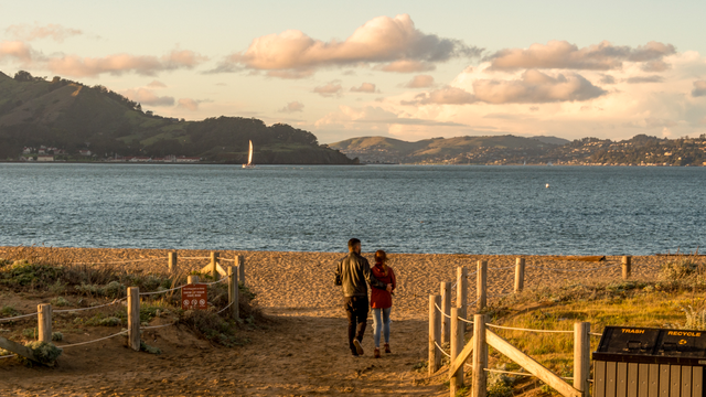 Visitors step out onto Crissy Field East Beach where the sunset casts warm light over the Golden Gate and San Francisco bay.