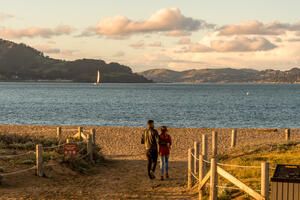 Visitors step out onto Crissy Field East Beach where the sunset casts warm light over the Golden Gate and San Francisco bay.