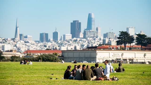 Park visitors enjoy a picnic sitting on the lawn at Crissy Field amongst the San Francisco city skyline on a sunny day.