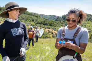 Two women carrying bug nets in a grassy field inspect specimen vials