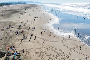 Aerial view of beach goers gathering around sand art at Ocean Beach.
