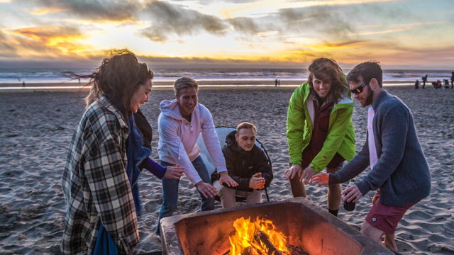 Ocean Beach visitors warm up beside a bonfire in the iconic fire pits as a golden sunset illuminates the sky.