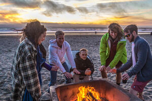 Ocean Beach visitors warm up beside a bonfire in the iconic fire pits as a golden sunset illuminates the sky.