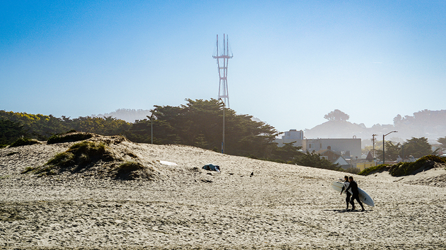 Surfers walk along Ocean Beach with Sutro Tower in the background