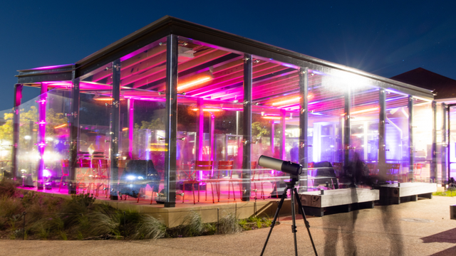 Long exposure photo of an enclosed outdoor picnic area in the Presidio glows with bright pink lights with an astronomy telescope seen in the foreground