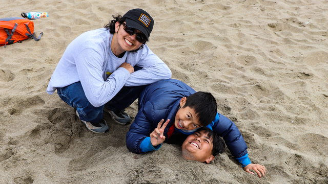 Three smiling and laughing youths posing at Baker Beach, one is buried in the sand with their head revealed