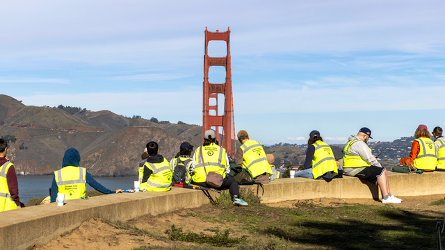 A group of volunteers in bright vests sit on a presidio bench with the golden gate bridge and marin headlands in view.