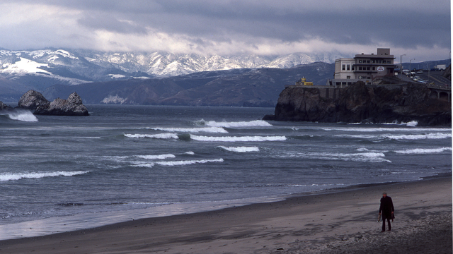Person walking along Ocean Beach, in front of Cliff House, with snowy Marin Headlands in Background