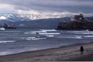 Person walking along Ocean Beach, in front of Cliff House, with snowy Marin Headlands in Background