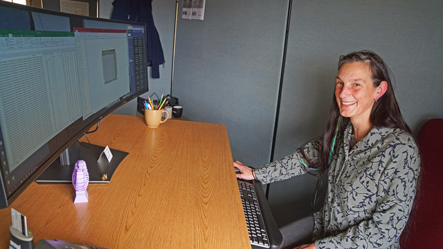 Lizzy Edson smiles behind a large curved computer monitor.
