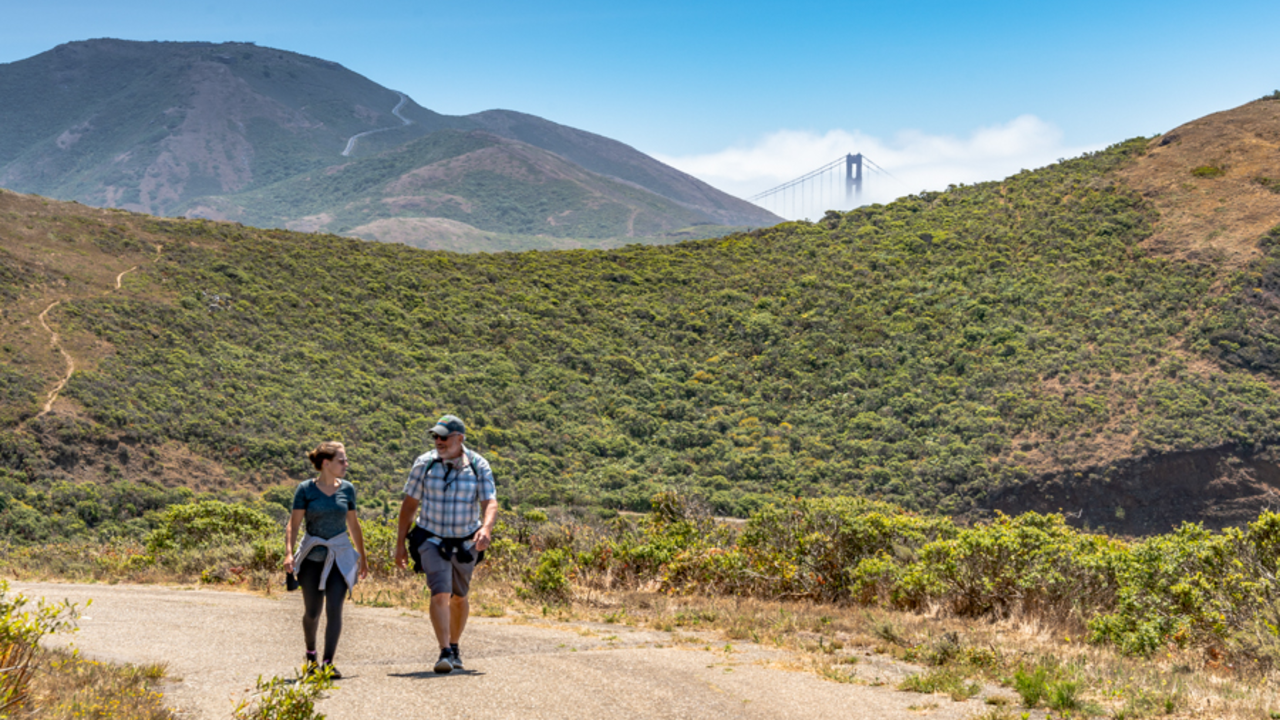 A couple hiking a dirt trail in the Marin Headlands. A Golden Gate Bridge tower can be seen peeking through the green mountain range in the background.