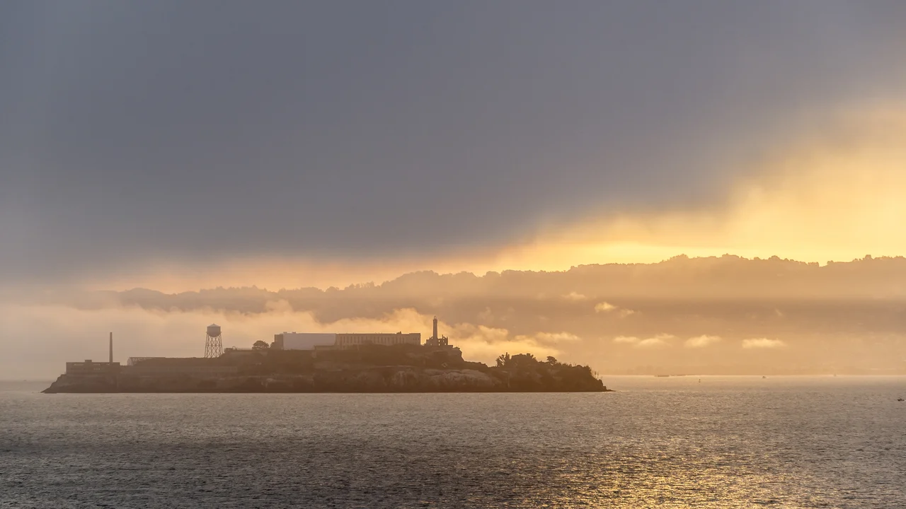 view of Alcatraz island with dark clouds above and a sunrise peeking through the clouds on the horizon