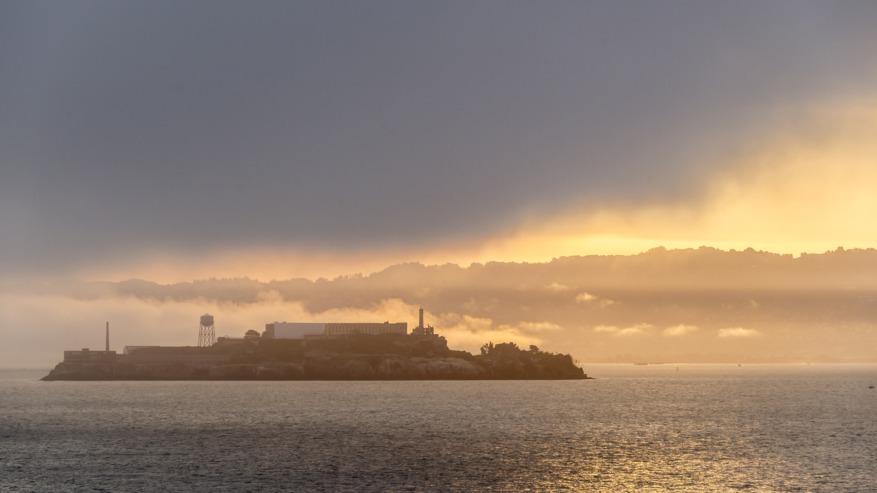 view of Alcatraz island with dark clouds above and a sunrise peeking through the clouds on the horizon