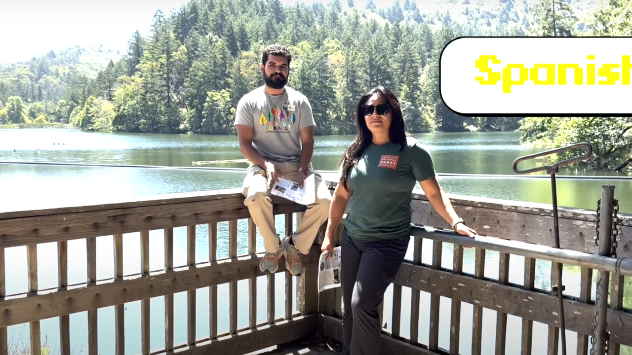 Parks Conservancy staff members sit on a fence in front of Lake Lagunitas, smiling at the camera. The title "SPANISH" is in the upper right.