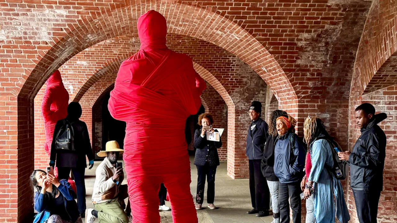 A group of Rafiki Coalition participants reacting with awe at a large human shaped sculpture wrapped in red gauze, a part of the Black Gold exhibit at Fort Point National Historic Monument in San Francisco.