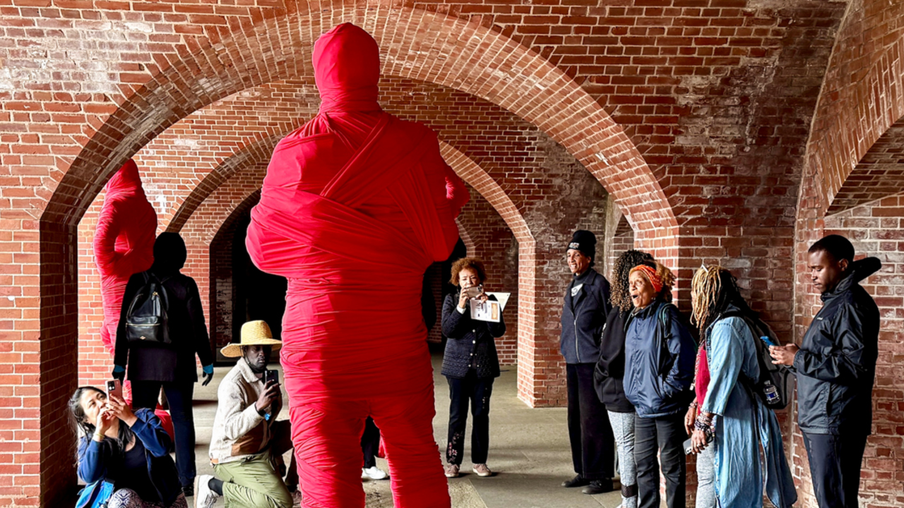 A group of Rafiki Coalition participants reacting with awe at a large human shaped sculpture wrapped in red gauze, a part of the Black Gold exhibit at Fort Point National Historic Monument in San Francisco.