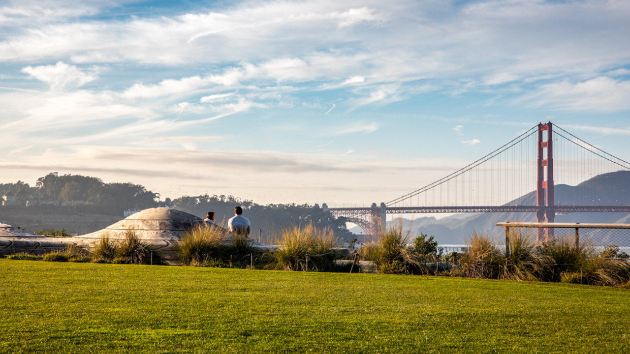A scenic view of the Golden Gate Bridge seen from the Presidio Tunnel Tops in San Francisco, visitors seen sitting on the iconic wooden park bench.