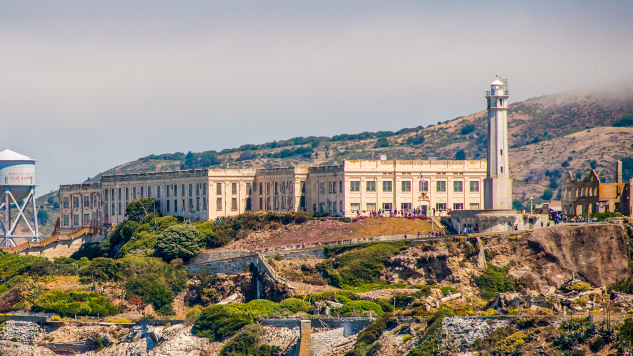 A scenic shot of Alcatraz Island from a distance. In view is the watertower, cellhouse, lighthouse, and other various buildings on the island.