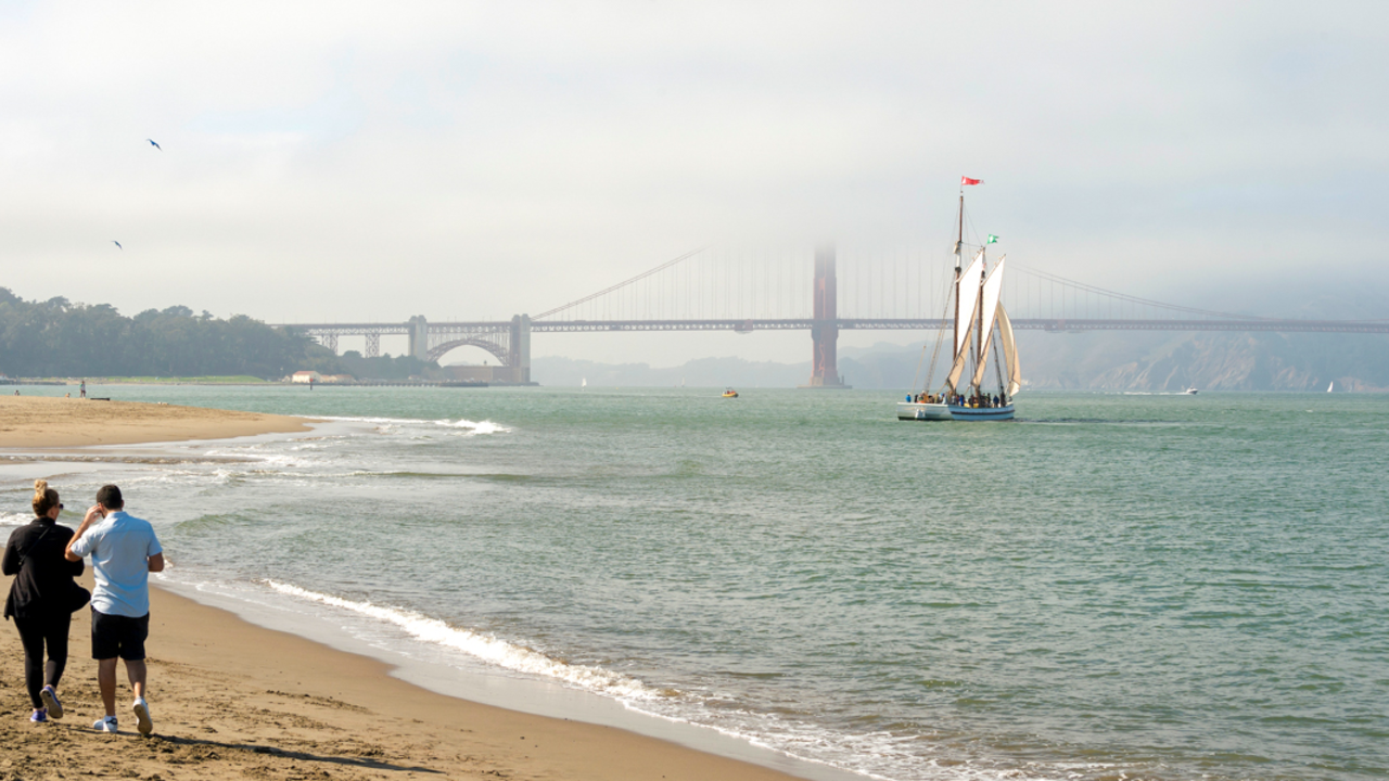 The sun breaks through a hazy day at Crissy Field East Beach, two park goers walk along the sand and a large sailboat can be seen on the water before the Golden Gate Bridge backdrop.