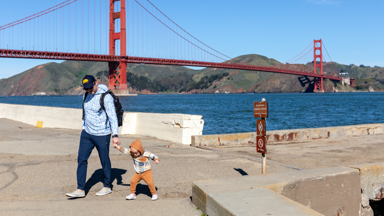 A parent and toddler walking hand in hand by the waterfront near the Warming Hut at Crissy Field. The Golden Gate Bridge is seen in the background.