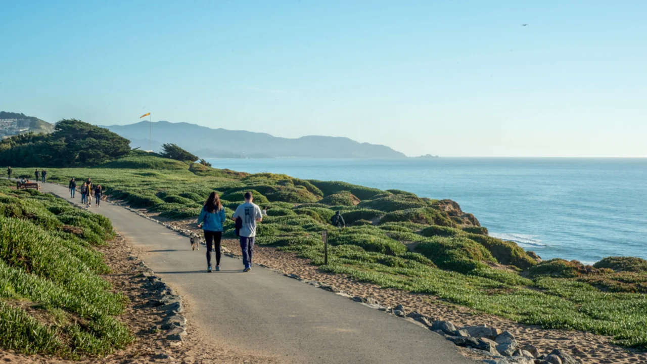 A view from Fort Funston in San Francisco. A paved trail leads down the coast, the bluffs give way to views of the mountain range and Pacific Ocean.