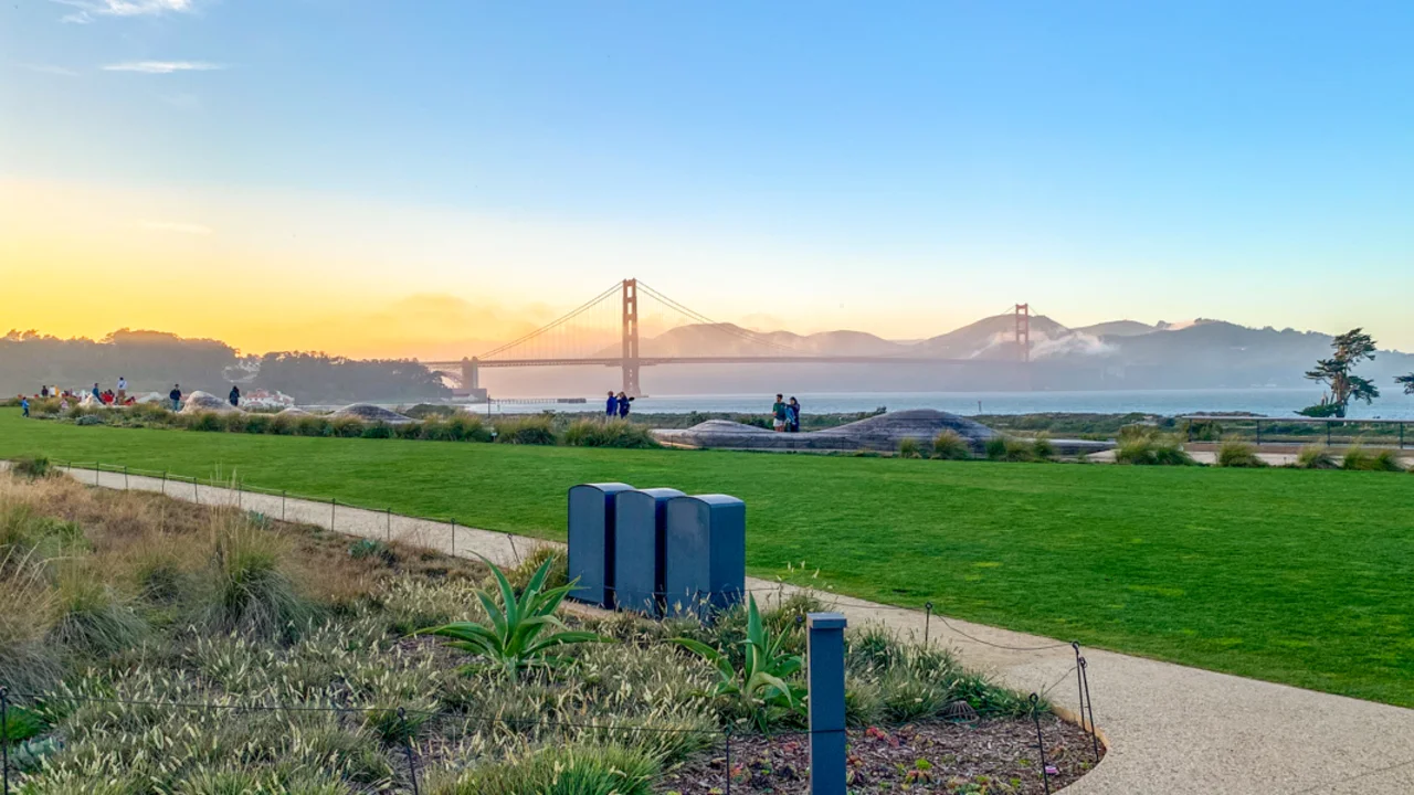 The verdant lawn at the Meadows of the Presidio Tunnel Tops. Park visitors are seen recreating about the park. The golden gate bridge is seen in the background.