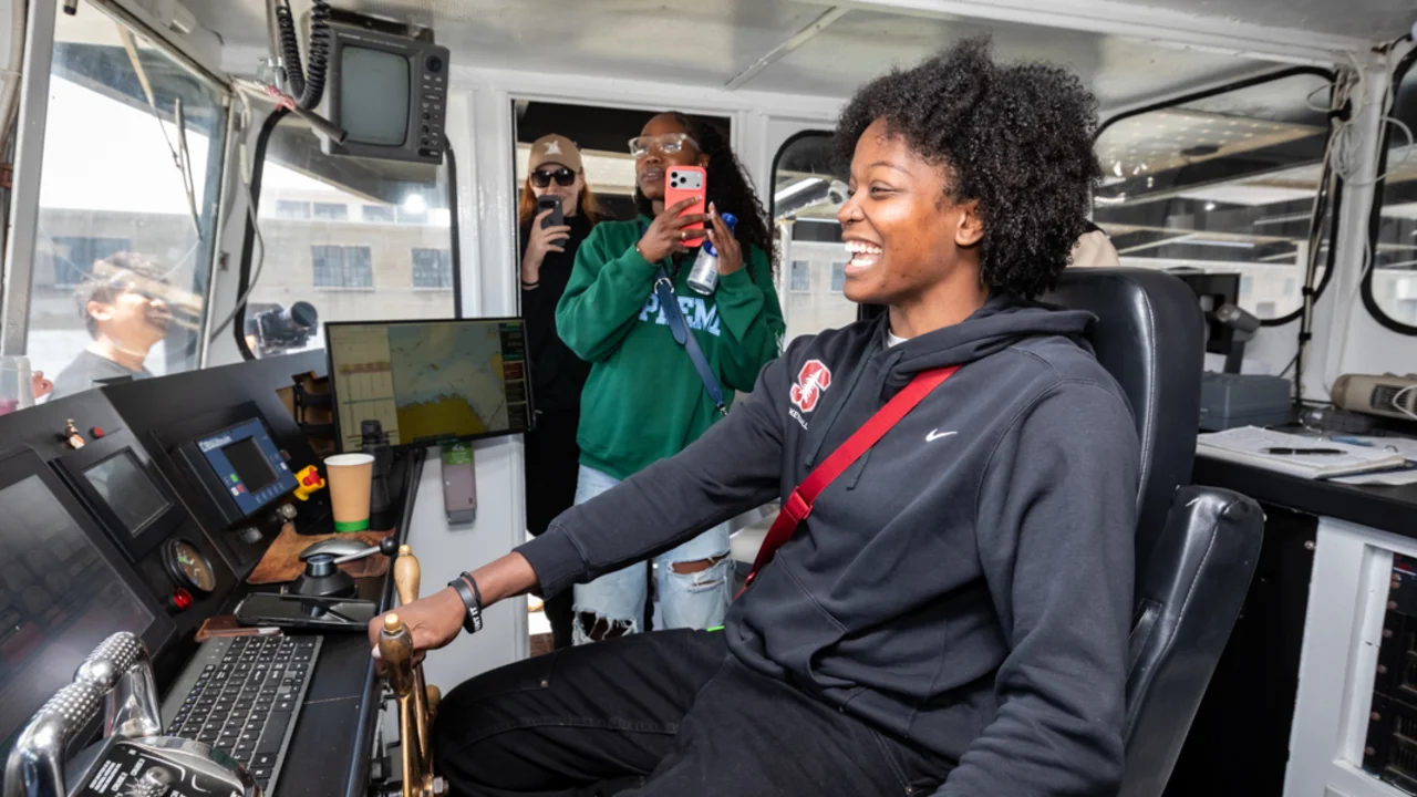A bay area basketball player smiles while pretending to operate the helm of an Alcatraz City Cruises ferry, with others observing and capturing the moment from behind.