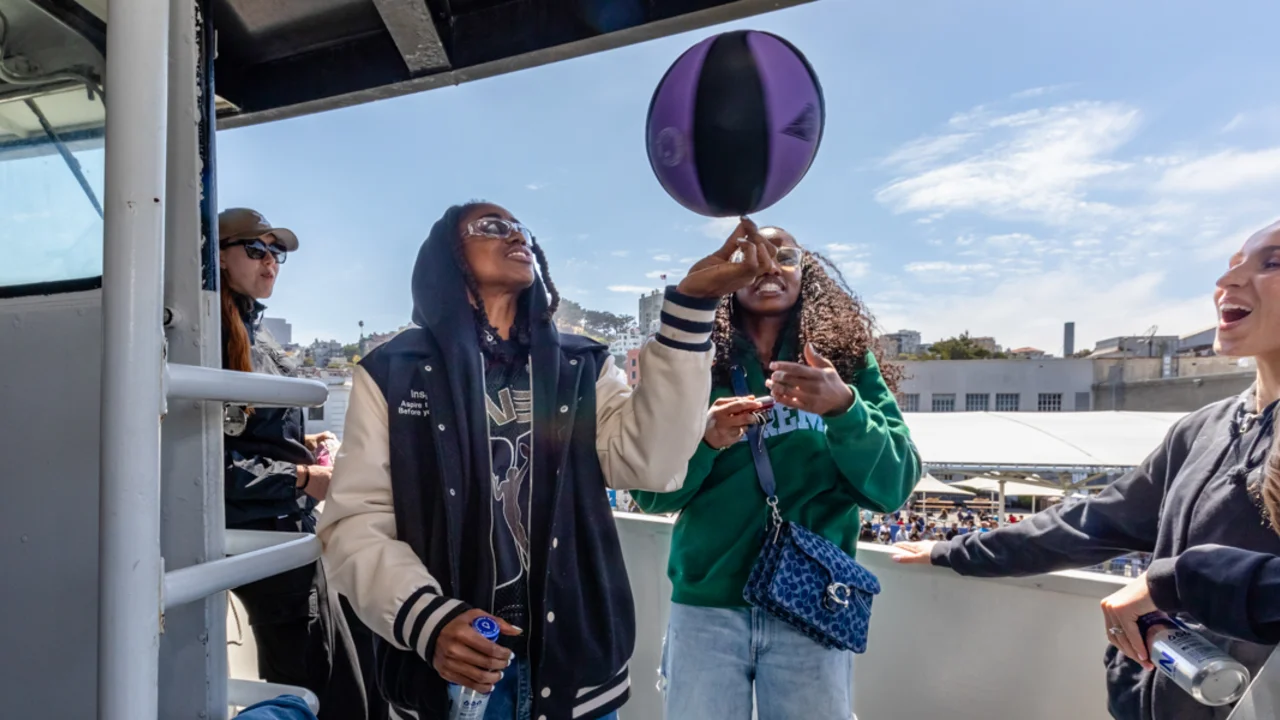 Bay area basketball players on a Alcatraz City Cruises ferry deck enjoy a sunny day, with one person balancing a purple and black ball on their finger while others laugh and look on.