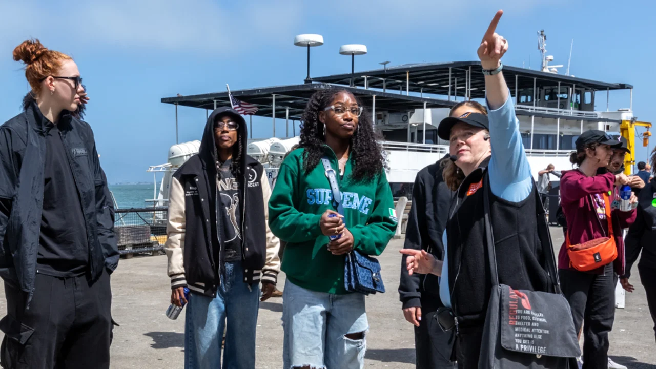A group of people stands on a dock near a ferry, with one person pointing upward toward the sky.